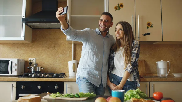 Молодая счастливая пара делает селфи фото во время приготовления завтрака на кухне дома — стоковое фото