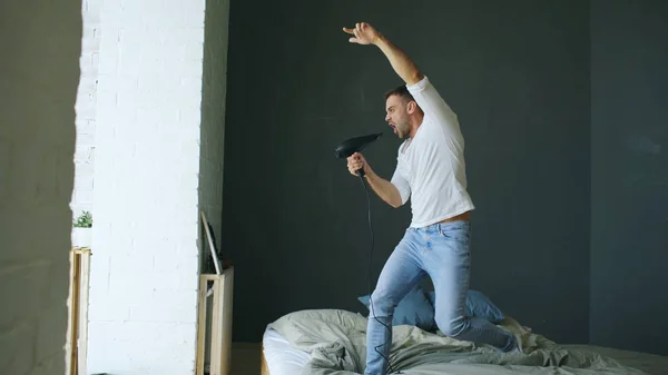A fiatalember éneklés haj szárító és táncoló rocknroll ágyon a hálószobában — Stock Fotó
