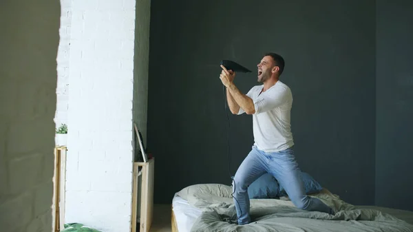 Joven cantando a secador de pelo y bailando rocknroll en la cama en el dormitorio — Foto de Stock