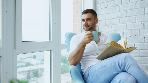 Молодий чоловік читає книгу, сидячи на балконі в сучасній квартирі — стокове фото