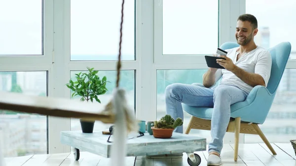 Vonzó férfi ül a széken a tetőtéri modern lakás erkéllyel felszerelt, digitális tabletta — Stock Fotó