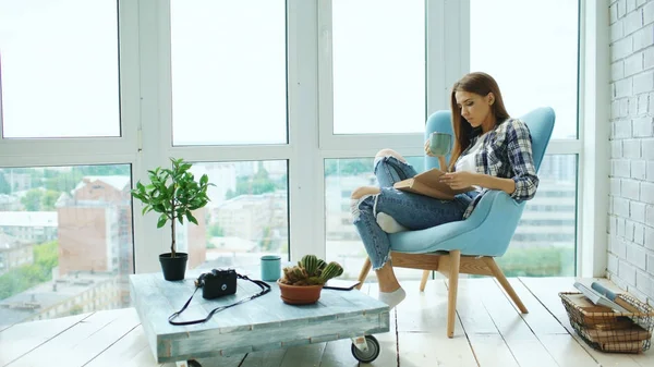 Fiatal, vonzó nő olvassa el a könyvet, és ital kávé ül a modern loft apartman erkéllyel — Stock Fotó
