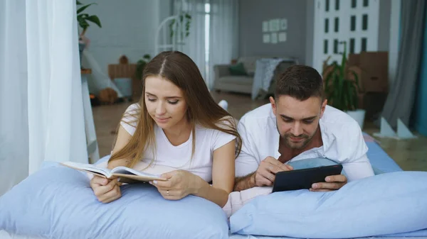 Красивый молодой человек, использующий планшетный компьютер, показывает фотографии на планшетном компьютере своей подруге, читающей книгу и лежащей в постели дома — стоковое фото