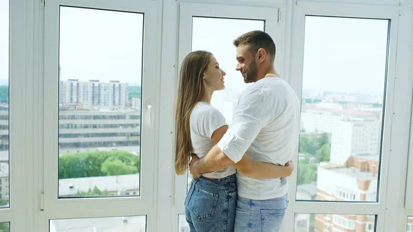 Giovane coppia felice abbracciando in piedi vicino alla finestra e godendo della vista dal nuovo appartamento — Foto Stock