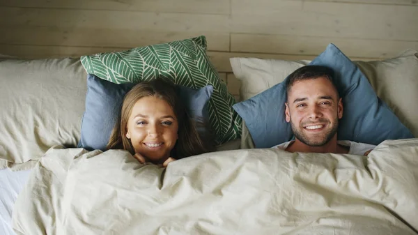 Κάτοψη του χαμογελώντας ζευγάρι διασκεδάζουν στο κρεβάτι κρύβονται κάτω από την κουβέρτα και να αναζητούν σε κάμερα — Φωτογραφία Αρχείου