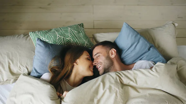Top view of couple having fun in bed lying under blanket looking into camera and kissing at home — Stock Photo, Image