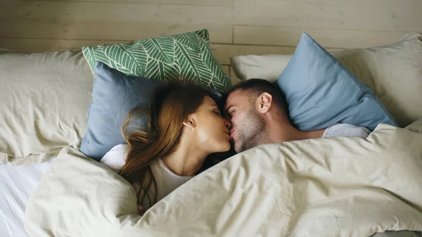 Вид на пару, развлекающуюся в постели, лежащую под одеялом, смотрящую в камеру и целующуюся дома — стоковое фото