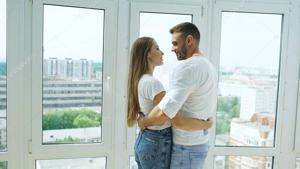 Young happy couple embracing standing near window and enjoying view from new apartment