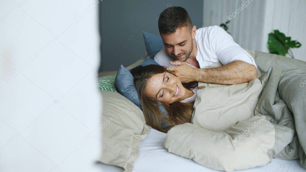 Young beautiful and loving couple wake up at the morning. Attractive man kiss and hug his wife in bed