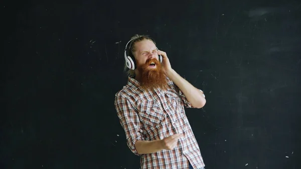 Retrato de barbudo jovem coloca em fones de ouvido e dança enquanto ouve música em fundo preto — Fotografia de Stock