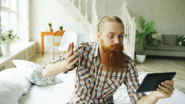 Mladý vousatý muž používající počítač tabletu s video chat seděl v posteli doma — Stock fotografie