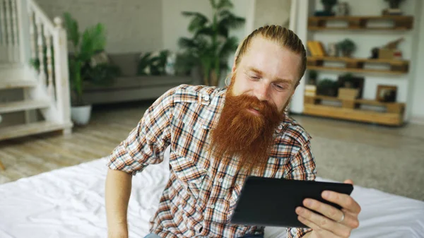 Fiatal szakállas férfi ült az ágyban otthon videochat tabletta számítógép — Stock Fotó