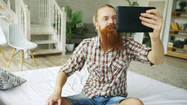 Jeune homme barbu à l'aide d'une tablette ayant chat vidéo assis dans le lit à la maison — Photo