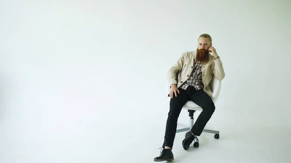 Grave uomo barbuto seduto sulla sedia girevole ufficio e guardando nella fotocamera su sfondo bianco — Foto Stock