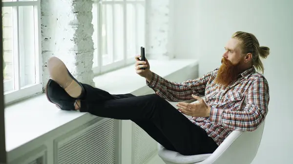 Jeune homme d'affaires barbu hipster parler chat vidéo sur smartphone tout en étant assis dans une chaise offce avec ses jambes sur le rebord de la fenêtre — Photo