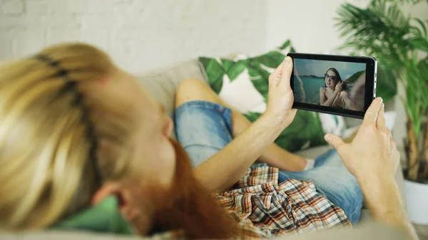 Young bearded man chatting with girlfriend using tablet computer lying in sofa at home — Stock Photo, Image