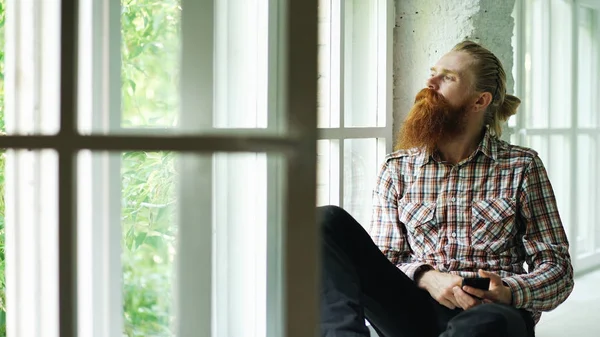 Szomorú szakállas férfi sms textil használ smartphone sit on ablakpárkányon otthon — Stock Fotó