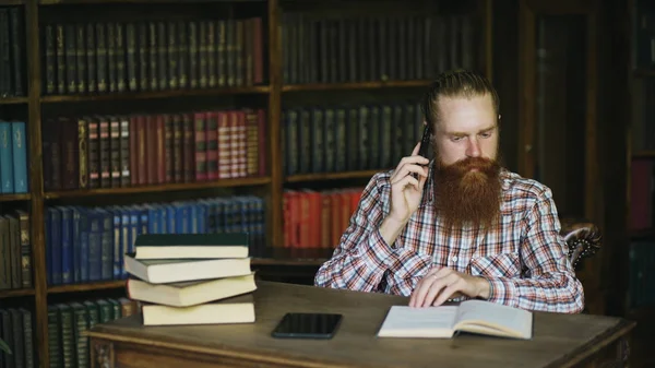 Jovem barbudo estudante falando telefone enquanto se prepara para exames na biblioteca da Universidade — Fotografia de Stock