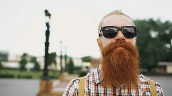 Retrato de hipster barbudo turista em óculos de sol olhando para a câmera e sorrindo para o fundo da cidade — Fotografia de Stock