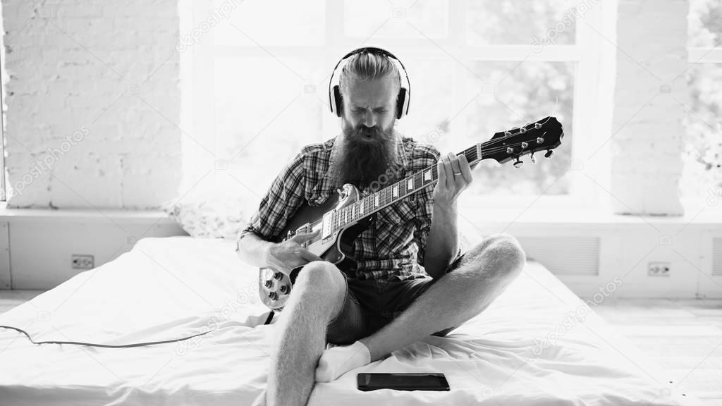 Attractive bearded man in headphones sitting on bed learning to play guitar using tablet computer in modern bedroom