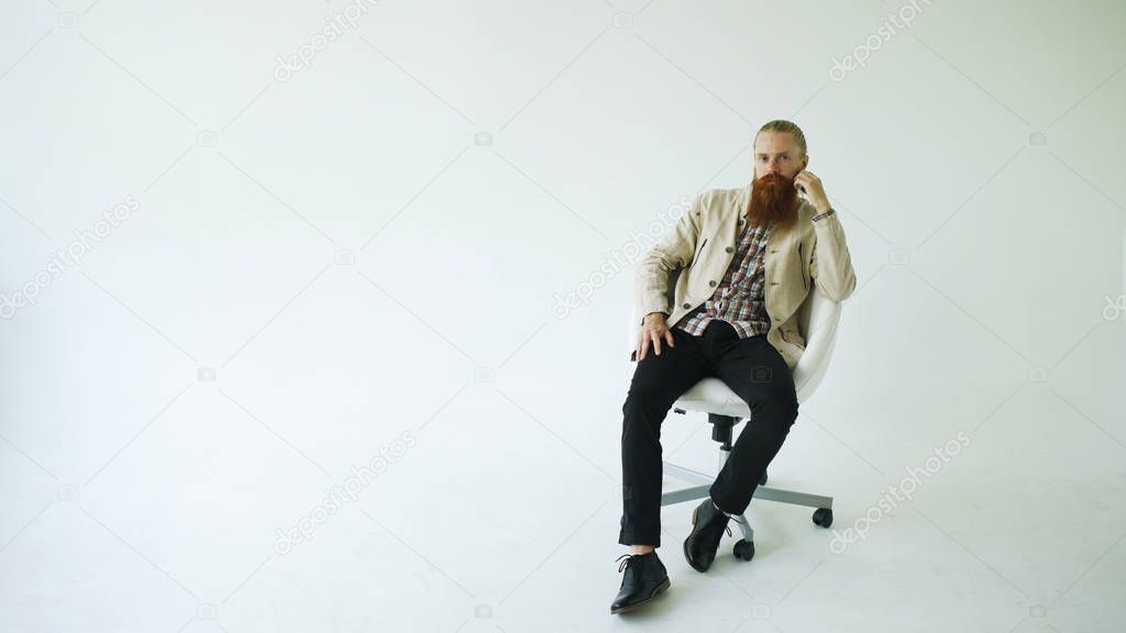 Serious bearded man sitting on swivel office chair and looking into camera on white background