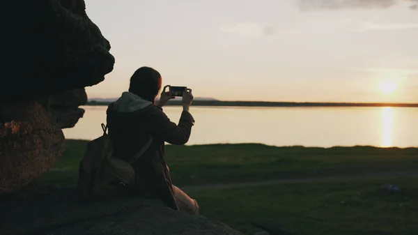 Молодая туристка-туристка фотографирует пейзаж на камеру смартфона после похода по скале на закате — стоковое фото
