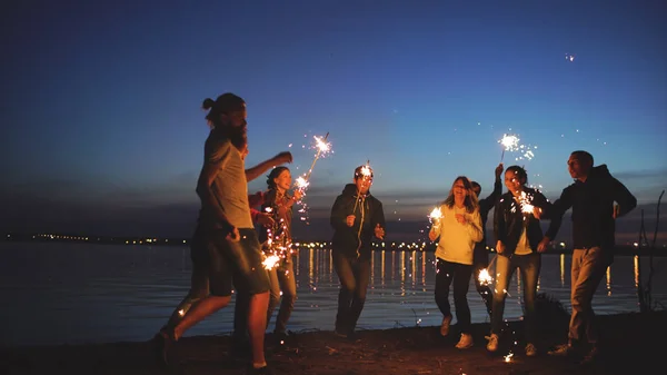 Group of young friends having a beach party. Friends dancing and celebrating with sparklers in twilight sunset — Stock Photo, Image