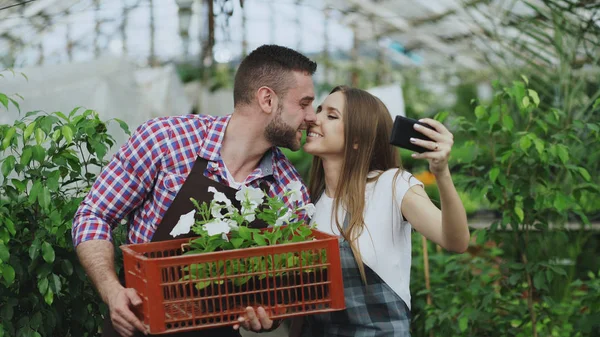 Alegre pareja cariñosa jardineros tomando foto selfie en la cámara del teléfono inteligente y besándose mientras trabaja en invernadero — Foto de Stock