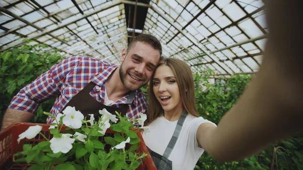 Wesoły kochający ogrodników para biorąc zdjęcie selfie na smartfonie aparatu podczas pracy w szklarni — Zdjęcie stockowe