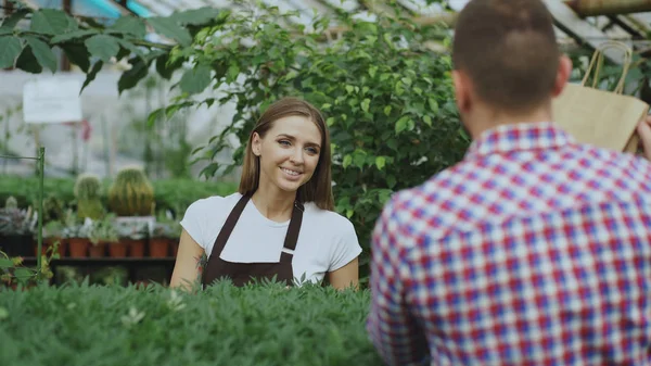 Jonge lachende verkoper bloemist werken in tuincentrum. Vrouw geeft boodschappentas aan klant op te slaan — Stockfoto
