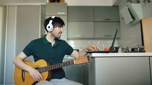 Veselý mladý muž se sluchátky na uších sedí v kuchyni, naučit se hrát na kytaru doma pomocí přenosného počítače — Stock fotografie