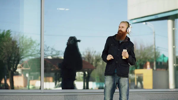 Feliz joven barbudo hipster hombre con auriculares y teléfono inteligente escuchar música y bailar en la calle de la ciudad — Foto de Stock