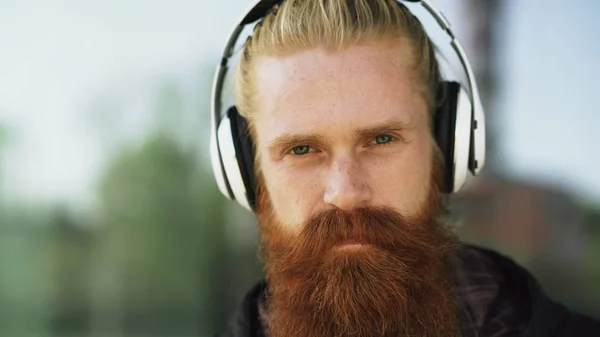 Retrato de close-up do jovem hipster barbudo com fones de ouvido ouvir música e sorrindo na rua da cidade — Fotografia de Stock