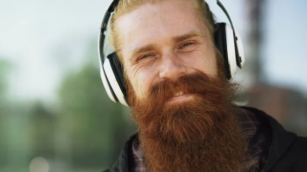 Närbild porträtt av unga skäggiga hipster man med hörlurar Lyssna på musik och ler mot gata — Stockfoto