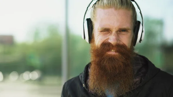 Närbild porträtt av unga skäggiga hipster man med hörlurar Lyssna på musik och ler mot gata — Stockfoto