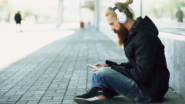 Jeune homme barbu hipster avec écouteurs assis sur la route et utilisant un smartphone pour écouter de la musique et surfer sur Internet en plein air dans la rue — Photo