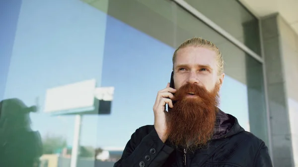Fiatal szakállas csípő ember koncentrált beszél telefonon, a citystreet és beszélgetés közelében irodaház — Stock Fotó