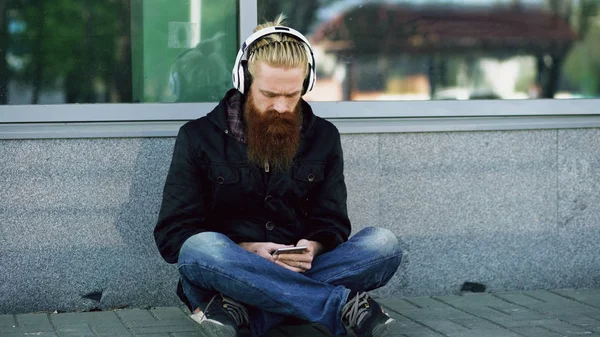 Fiatal szakállas csípő ember ül a közúti fejhallgató, és használ a smartphone hallgatni zene és internet szörfözés szabadban utca — Stock Fotó