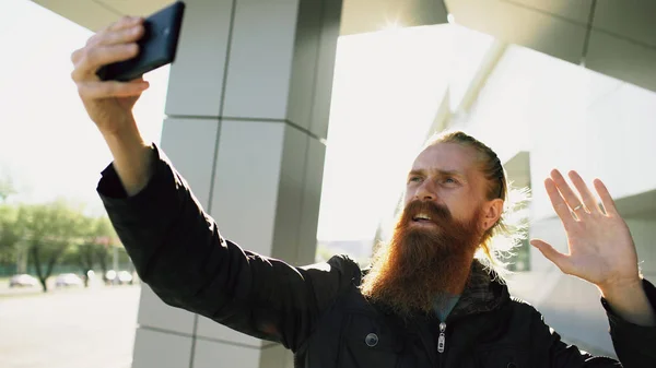 Jovem homem hipster barbudo ter bate-papo de vídeo on-line com câmera do smartphone enquanto viaja rua da cidade — Fotografia de Stock