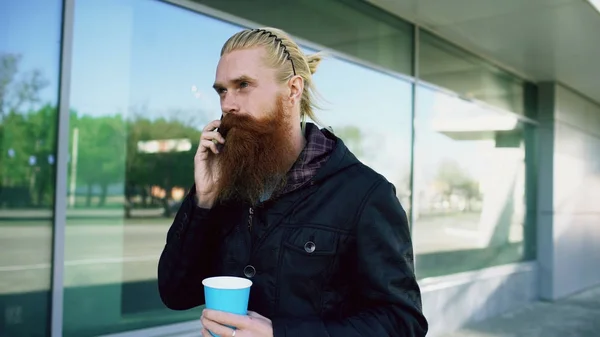 Boldog szakáll fiatalember beszél a smartphone és a kávéfogyasztás, miközben sétál az utcán — Stock Fotó