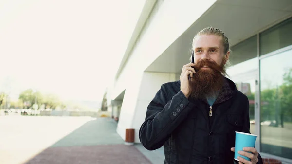 Glad skägg ung man talar på smartphone och dricker kaffe medan promenader i gatan — Stockfoto