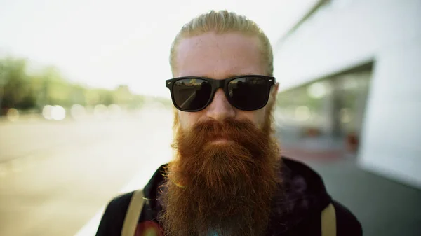 Vértes portré fiatal szakállas csípő férfi napszemüveg, mosolyogva és pózolt utazás a város utca — Stock Fotó