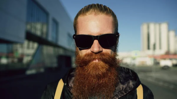 Retrato de close-up de jovem homem hipster barbudo em óculos de sol sorrindo e posando enquanto viaja pela rua da cidade — Fotografia de Stock