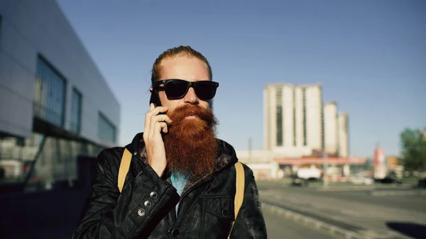 Junger bärtiger Hipster mit Sonnenbrille lächelt und spricht Smartphone, während er in der Stadt unterwegs ist — Stockfoto