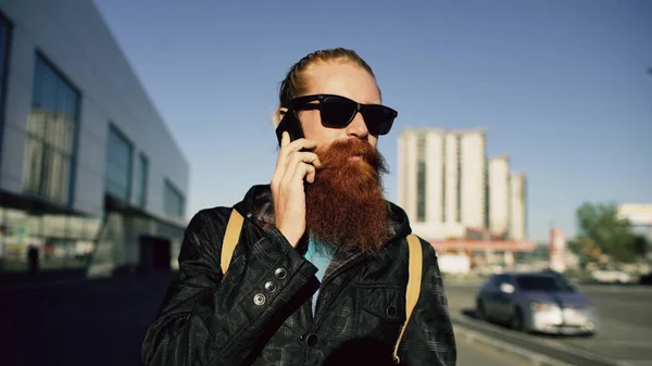 Jovem homem hipster barbudo em óculos de sol sorrindo e falando smartphone enquanto viaja rua da cidade — Fotografia de Stock