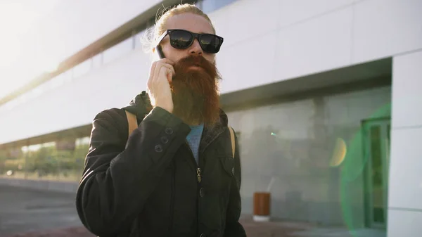Młody Brodaty mężczyzna hipster w okulary, uśmiechając się i mówienie smartfona podczas podróży miasta ulicy — Zdjęcie stockowe