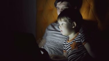 Genç Baba ve küçük oğlu tablet bilgisayar kullanarak ve evde akşam yatakta yatan süre konuşuyor karikatür film izlerken
