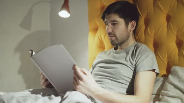 Молодой отец читает книгу, в то время как его маленький сын прыгает на кровать перед сном — стоковое видео