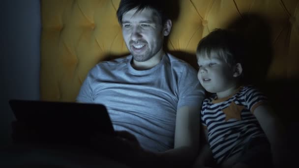 Joven padre y su pequeño hijo viendo películas de dibujos animados con computadora portátil mientras está acostado en la cama por la noche en casa — Vídeo de stock