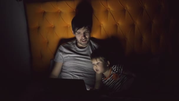 Νεαρό πατέρα και τον μικρό γιο του βλέποντας ταινία κινουμένων σχεδίων, χρησιμοποιώντας τον υπολογιστή tablet και μιλάμε ξαπλωμένοι στο κρεβάτι το βράδυ στο σπίτι — Αρχείο Βίντεο